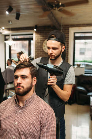 Stylist making modern hairdo for young man. Successful businessman sitting at barber shop and dry hair. Style, beauty, modern life conceptの写真素材