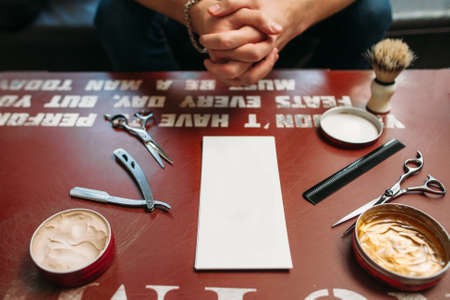 Blank card with barber tools on table free space. Side view on gules workplace with hairstyling instruments and hands of sitting man on background, Work, barbershop, manhood conceptの写真素材