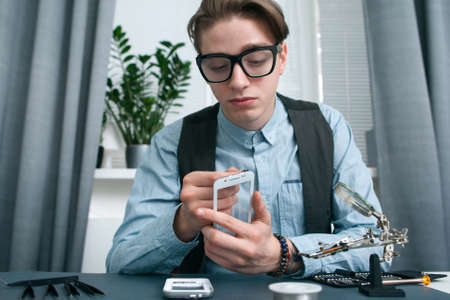 Young man seriously looking at smartphone screen. Geek in glasses intently examing disassembled mobile phone in office, free space. Electronic fixing, modern technology, business conceptの写真素材