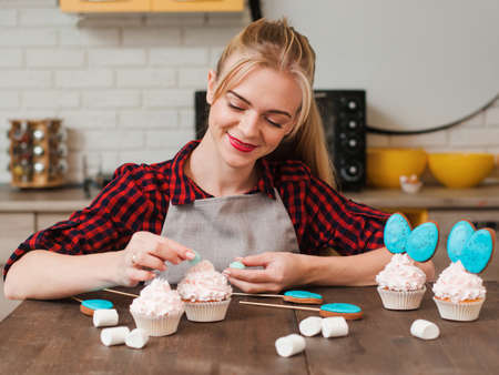 Girl decorating cup cakes with white cream and blue cake-pops at kitchen wooden table. Culinary masterclass. Easter gift, small business, delivery of sweets, craftmanship conceptの写真素材