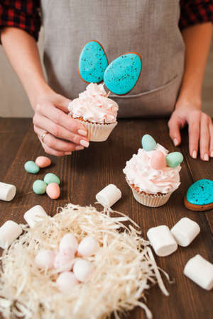Decorated cup cakes and nest with small white eggs for easter celebration on wooden table. Holiday gift, small business, delivery of sweets, craftmanship conceptの写真素材