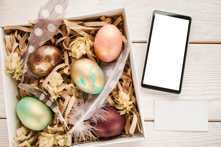 Gift easter box with five color eggs, feathers and flowers on white wood table. Smartphone with...