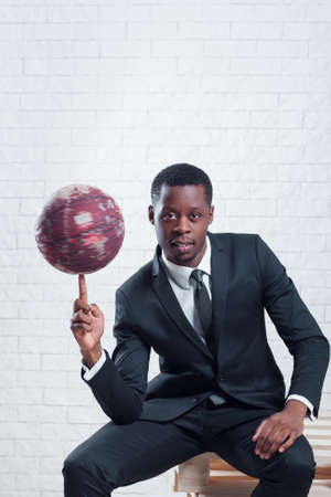 Successful african sportsman with a basketball wearing business suit on white brick wall background.の写真素材