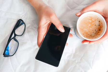 Unrecognizable business woman morning start. Hand hold cup of fresh coffee and mobile phone with black screen, close up. Messaging, reading news, e-mail concept.の写真素材