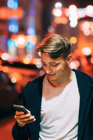 Handsome young man using smartphone while walking in night city streets. Smiling guy reading message outdoorの写真素材