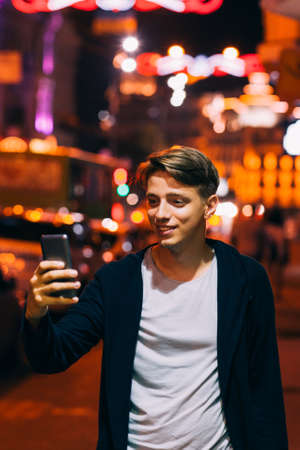 Young man take photo on smartphone while walking in night city streets. Smiling guy on blurred lights backgroundの写真素材