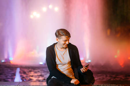 Handsome young man using smartphone while walking in night city streets. Smiling guy reading message outdoorの写真素材
