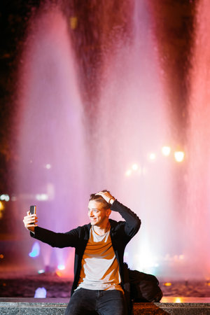 Young man take selfie on smartphone while walking in night city streets. Smiling guy on blurred lights backgroundの写真素材
