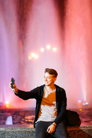 Young man take selfie on smartphone while walking in night city streets. Smiling guy on blurred...