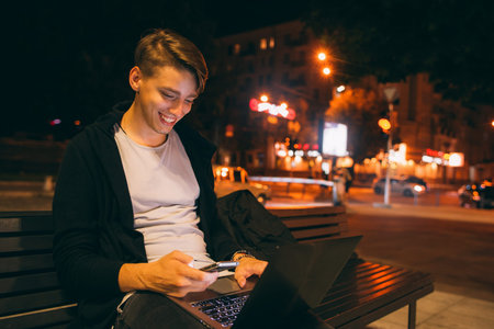 Young happy freelancer working in city street in night time, blurred lights background, free space....