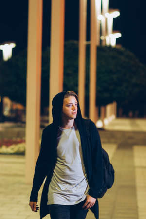 Young handsome casual student in hood outdoors. Portrait of hooded guy walking through night cityの写真素材