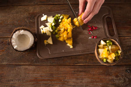 Preparing of tropical fruit salad in coconut shell. Orange, apple, kiwifruit and pomegranate ingredients are cut on brown wooden deskの写真素材