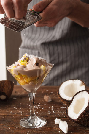 Culinary art of fresh fruit salad in martini glass with coconut flakes. Unrecognizable man makes a sweet dessert with decorationの写真素材