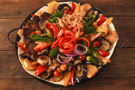 Dish with stewed meat, different kind of vegetables, field mushrooms, laid on lavash and big copper plate, and decorated with sliced onion and pepper. Top view picture on wooden tableの写真素材