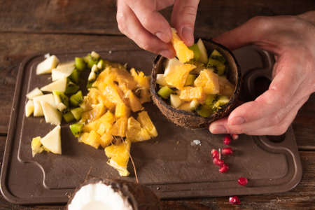 Close up process of preparing and decoration of tropical fruit salad in coconut shell. Orange, apple, kiwifruit and pomegranate ingredients are mixed in nut bowlの写真素材