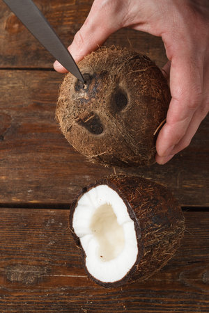 Close up coconut in process of opening by knife. How to break a shell of tropical fruit. Brown wooden backgroundの写真素材