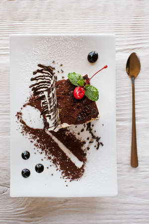 Tiramisu on white square cut plate with decoration from chocolate, cherry and mint. Sweet dessert serving in restaurant on wooden table, top viewの写真素材