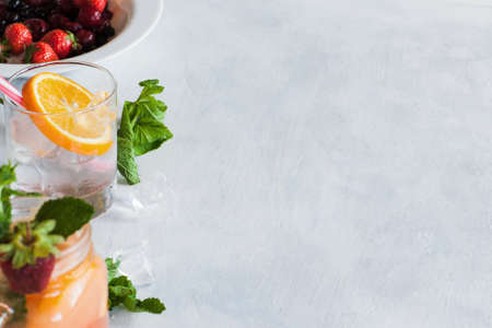 Background with fresh fruit cocktails preparing. Glass of cold drink with citrus slice, mint and ice, berries in bowl ready to use, refreshment and summer concept, close up picture with free spaceの写真素材