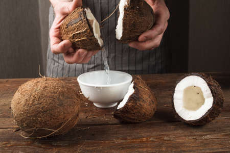 Fresh coconut water pouring into white bowl on wooden table. Preparing of beauty products, cosmetic oils and body lotions from organic ingredients of tropical fruitsの写真素材