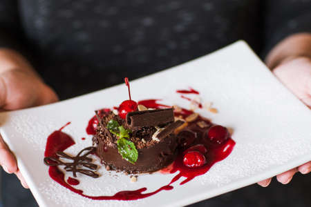 Customer holds chocolate cake with cherry jam, decorated by sweet red berry, almond and mint. Delicious dessert serving in restaurant, close up pictureの写真素材