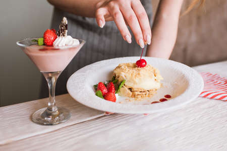 Process of decoration sweet desserts by culinary specialist in restaurant. Final touch on delicious napoleon cake with cherry and ready made fruit souffle nearby, free space belowの写真素材