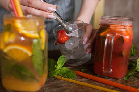 Waiter puts piece of ice into fruit cocktails. Fresh citrus and strawberry cold drink with mint on wooden table, concept of refreshment and satisfying thirst in summer, close up pictureの写真素材