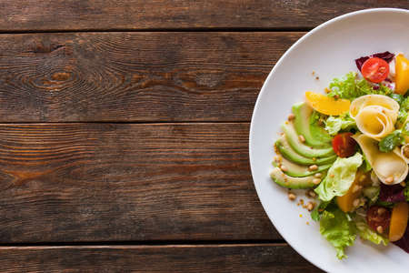 Lettuce salad with cheese and fruits on plate. Fresh vegetables decorated pine nuts, avocado, pearch and pear, serving in restaurant, top view with free space on leftの写真素材