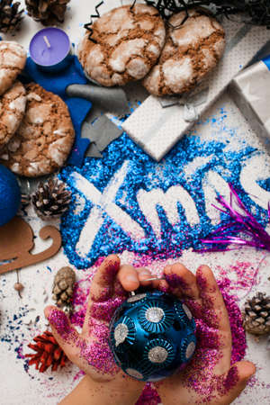 Christmas festive background reversed top view. Little kid holds ornament blue ball in purple glittering hands on xmas inscription and handmade decoration backdrop. Holiday and celebration conceptの写真素材