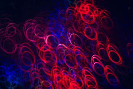 Blurry abstract picture of colorful ellipses in motion on black background. Bokeh of defocused curls, vivid neon blue and red leds, glowing festive backdrop, holidays and celebrationsの写真素材