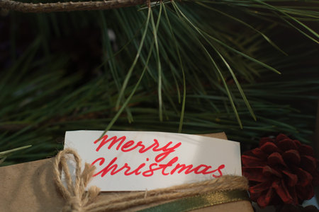 Festive background of Merry Christmas greeting. Close up handmade striped package with memo on pine branch backdrop. Presents, New Year traditions and congratulation conceptの写真素材