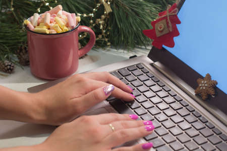 Chatting and Christmas congratulations. Woman typing on laptop near festive decorated pine and cup of marshmallows, close up picture. Greetings in social network, parents and friends abroad conceptの写真素材