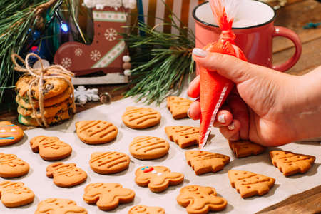 Icing process of Christmas bakery. Unrecognizable woman decorates homemade gingerbread cookies on cooking sheet, near pine branch with festive decor. Family culinary and New Year traditions conceptの写真素材