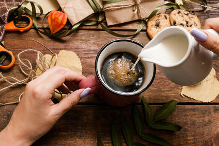 Autumn holidays and warm drinks. Top view unrecognizable woman pouring creams into cup of coffee on wooden table near small wrapped gifts, chocolate cookies, scissor and bandsの写真素材