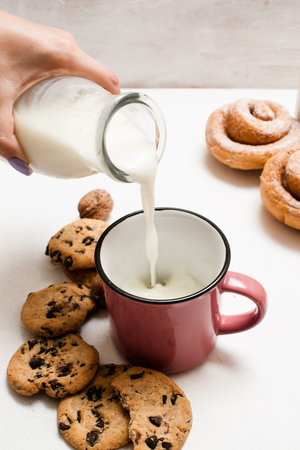 Pastry organic breakfast with milk and scones, close up. Woman pouring fresh lacto drink into cup near wholegrain cookies and baked rolls laying on white tableの写真素材