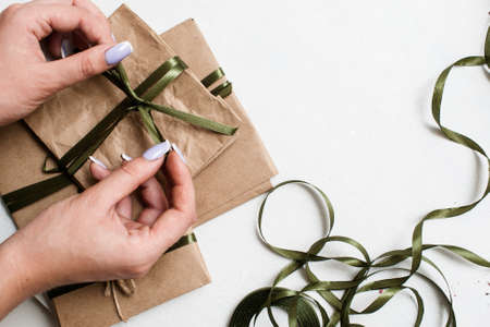 Holiday background of preparing eco presents. Small elegant gifts on white table with decorative ribbons are taped by unrecognizable woman, top view picture with free spaceの写真素材