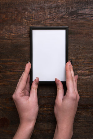 Woman hands holding white frame against wooden background. Hanging certificate or diplomaの写真素材