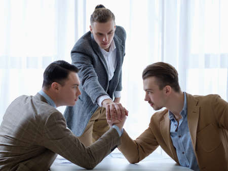 business competition. young man refereeing two rivals in their  arm wrestling fight. cruel corporate worldの写真素材
