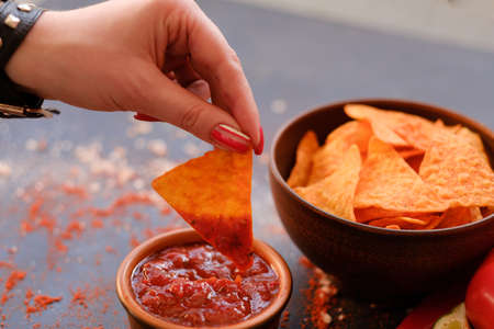 junk fast food and unhealthy eating. nacho tortilla chips in a bowl. woman hand dipping crunchy spicy triangular crisps in salsa tomato sauceの写真素材