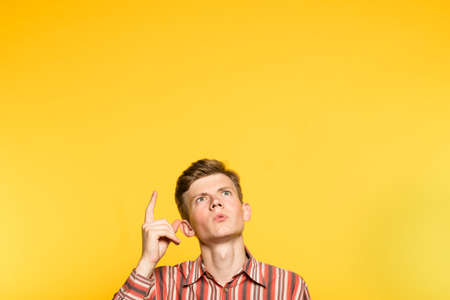 Amazed impressed astonished man looking and pointing up with a finger. portrait of a young guy on yellow background popping up or peeking out from the bottom. free space for advertisement.の写真素材