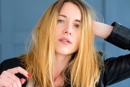 Disheveled sexy edgy young woman in black leather rocker jacket with ruffled hair. blond girl portrait on blue backgroundの写真素材