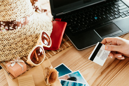 booking summer vacation trip online. traveling holidays and tourism concept. woman holding credit or debit bank card paying for the tour on the internet. laptop hat sunglasses on the desk.の写真素材