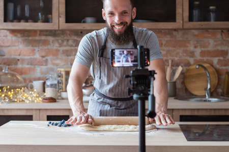 food blogger streaming live. bearded hipster man communicating with subscribers through phone camera. chef rolling out dough with a pin.の写真素材