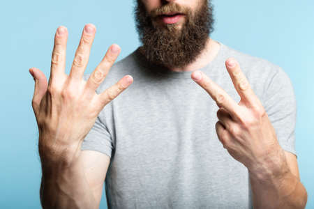 bearded man showing number seven with his hand. cropped shot of a male torso on blue background. casual hipster in grey t-shirt counting gesture.の写真素材