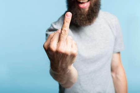 bearded man showing middle finger. provocation defiance attitude and offensive rude behavior concept. cropped shot of a male torso on blue background. casual hipster in grey t-shirt.の写真素材