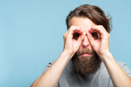 funny ludicrous joyful comic playful man pretending to look through binoculars made of hands. portrait of a young bearded guy on blue background. emotion facial expression conceptの写真素材
