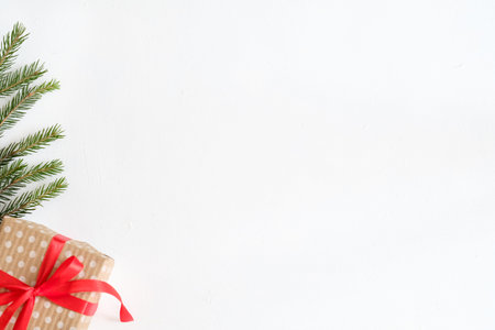 festive simple seasonal background. present wrapped in dotted craft paper and tied with red ribbon on white backdrop. handcrafted gift and fir tree branches decor. free space concept.の写真素材