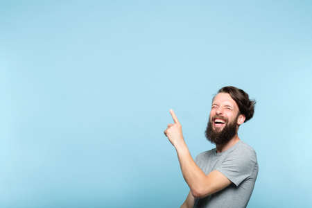 laughing man pointing behind sideways to a virtual object or text. copy space for advertisement or product placement. portrait of a bearded hipster guy on blue background.の写真素材