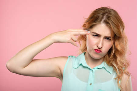 kill me please. bullet to the head. girl pointing a finger gun to her temple. emotional breakdown and stress concept. portrait of a beautiful girl on pink background.の写真素材