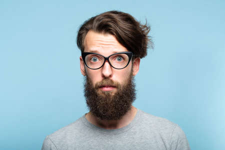 worried nervous alarmed bearded hipster guy wearing cat eye glasses. stylish modern fashionist. portrait of a geeky quirky eccentric man on blue background.の写真素材