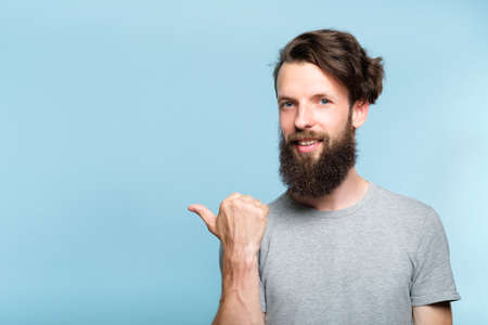 smiling young man pointing sideways with thumb as if showing smth. portrait of a bearded guy on blue background. copy space for advertisement.の写真素材